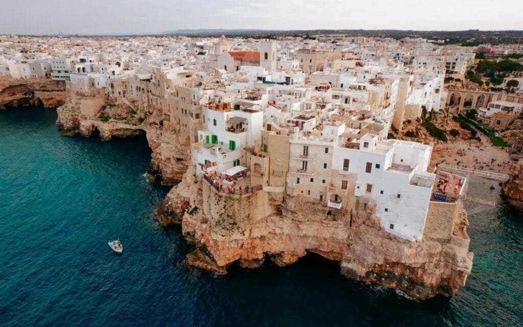 Aerial view of the coastal town of Polignano a Mare in Puglia, Italy, with whitewashed buildings perched dramatically on limestone cliffs above the Adriatic Sea.