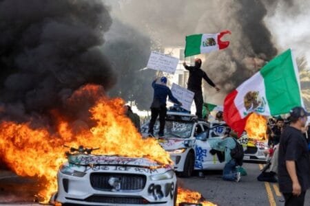 Demonstrators wave flags from a wrecked car, standing beside burning Waymo vehicles (Picture: Getty)